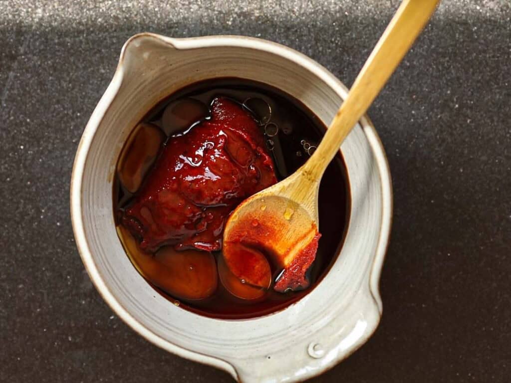 A ceramic bowl containing gochujang sauce. A wooden spoon rests inside, covered in the sauce. The bowl is placed on a dark, textured surface.